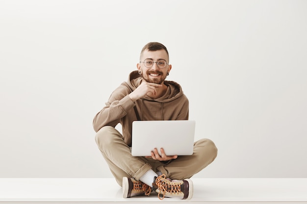 Hipster guy sitting with crossed legs with laptop and smiling pleased