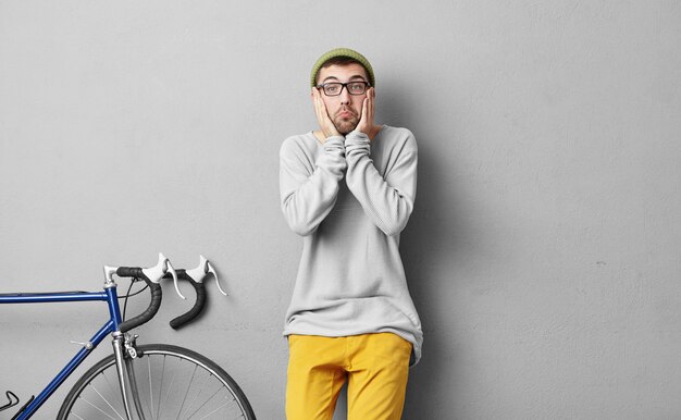 Hipster guy with surprised expression, wearing fashionable clothes, standing near his bicycle