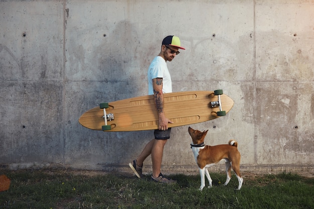 Hipster longboarder and a young brown and white basenji dog looking at one another next to a gray concrete wall