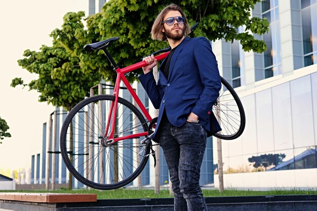 長いブロンドの髪を持つ流行に敏感な男性は、背景に高層ビルがある公園で彼の肩に赤いシングルスピード自転車を保持しています。