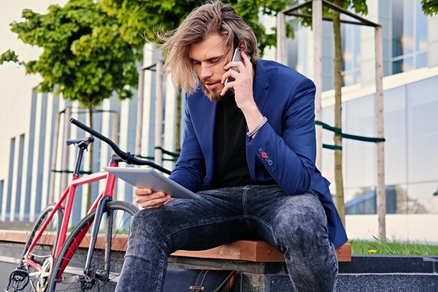 長いブロンドの髪を持つ流行に敏感な男性はスマートフォンで話し、バックグラウンドでシングルスピード自転車とタブレットPCを保持します。