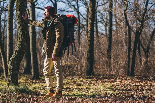 Hipster man traveling with backpack in autumn forest wearing warm jacket and hat, active tourist, exploring nature in cold season