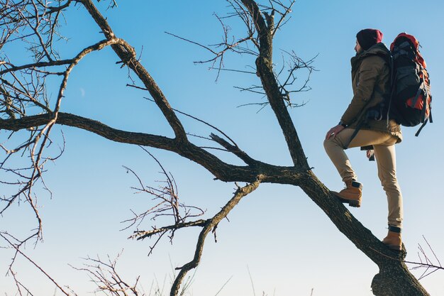 Hipster man traveling with backpack, standing on tree against sky, wearing warm jacket, active tourist, exploring nature in cold season