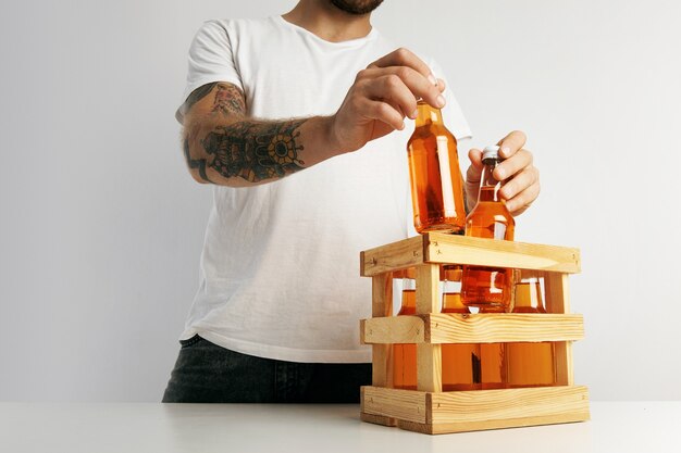白いテーブルの上の木製の箱にオレンジ色のレモネードのボトルを詰める無地の白いTシャツのヒップスター