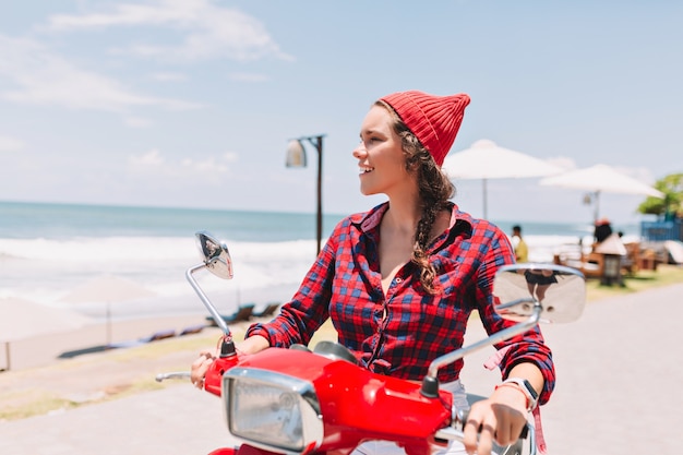 푸른 물과 바다의 배경에 햇빛에 빨간 자전거에 격자 무늬 셔츠와 빨간 모자 드라이브를 입은 소식통 예쁜 아가씨.