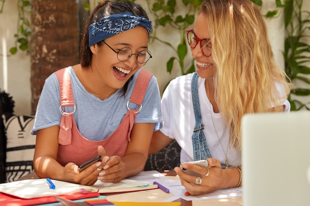 Hipster students search information on internet website, laugh happily as notice funny picture in mobile phone, pose together at desktop with laptop computer and notepad, enjoy communication