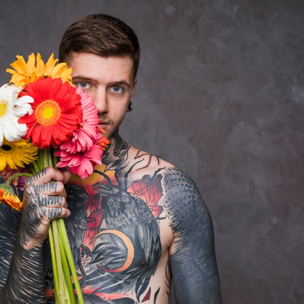 Hipster tattooed young man holding colorful gerbera flowers in hand standing against grey backdrop