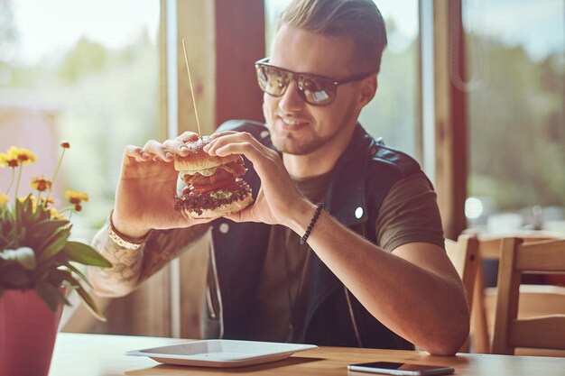 スタイリッシュなヘアカットとあごひげを生やしたヒップスターがテーブルに座り、道端のカフェでハンバーガーを食べて食事をすることにしました。