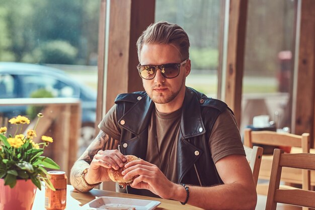 Hipster with a stylish haircut and beard sits at a table, decided to dine at a roadside cafe, eating a hamburger.