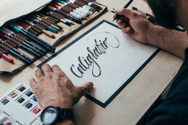 Молодой хипстерский художник-иллюстратор в простой черной футболке создает аутентичный и уникальный рисунок ручной надписи в своей яркой индустриальной студии