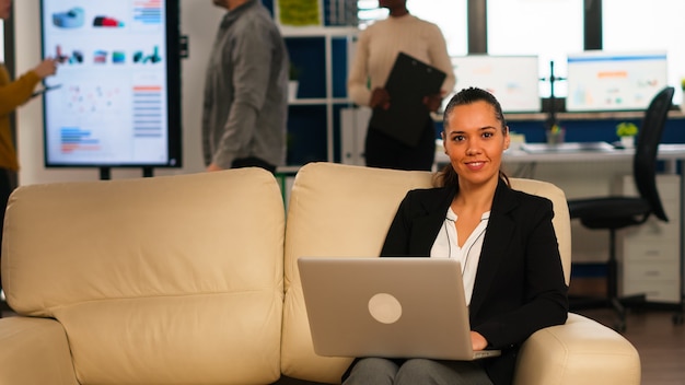 Hispanic business woman smiling at camera sitting on couch typing on computer while diverse colleagues working in background. Multiethnic coworkers analysing startup financial reports in modern office