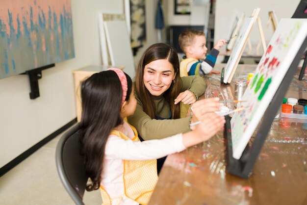 ヒスパニック系の女性インストラクターが笑顔で、子供向けの絵画教室でキャンバスに絵を描く方法を教えています