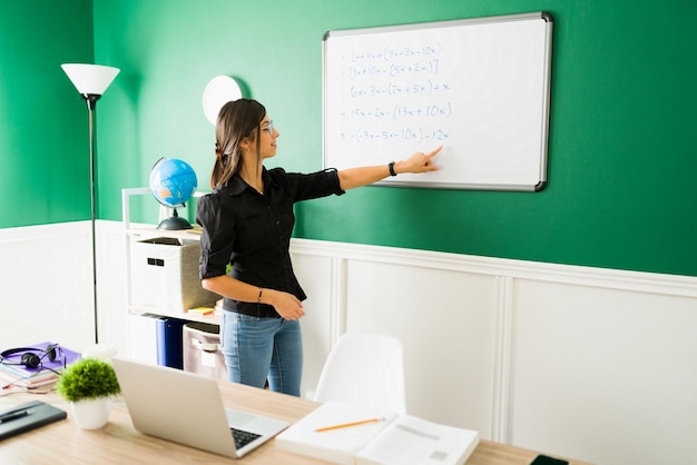 Hispanic female teacher giving a math lesson and writing equations on the blackboard during a virtual class