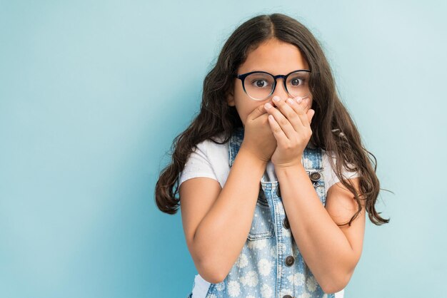 Hispanic girl in shock covering mouth with hands while making eye contact against plain background