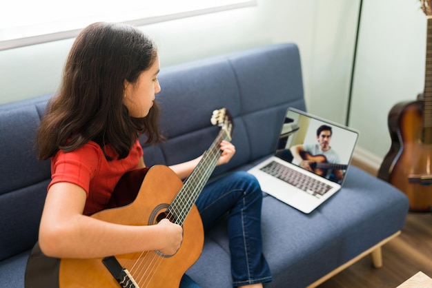 自宅のソファに座ってノートパソコンでビデオ通話中にギターの先生の指示を聞いている音楽スキルを持つヒスパニック系の女の子