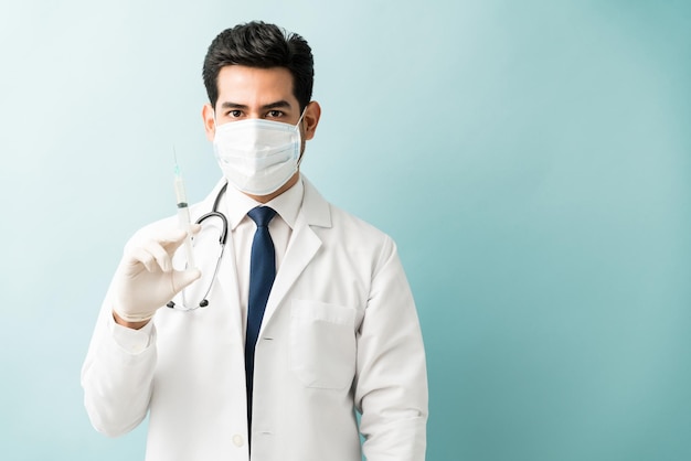 Free photo hispanic male doctor holding syringe while wearing mask against isolated background