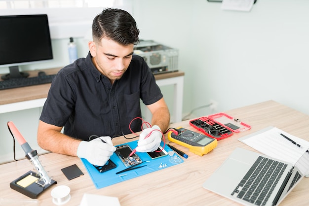 Ingegnere ispanico che controlla le connessioni di uno smartphone danneggiato con un multimetro presso la sua officina