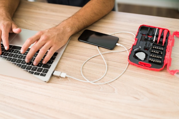 Hispanic male technician typing on laptop and connecting a smartphone to the computer to make a data backup at his repair shop