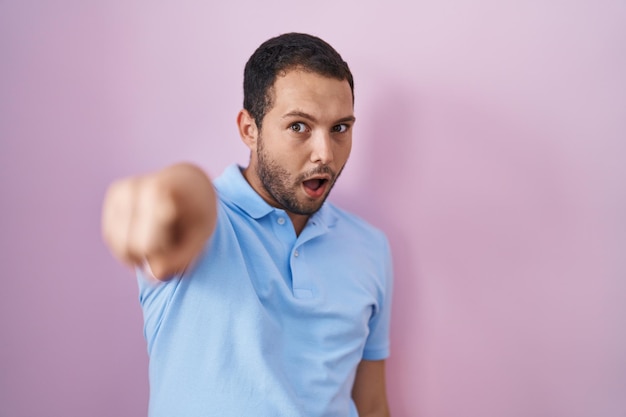 Hispanic man standing over pink background pointing with finger surprised ahead, open mouth amazed expression, something on the front