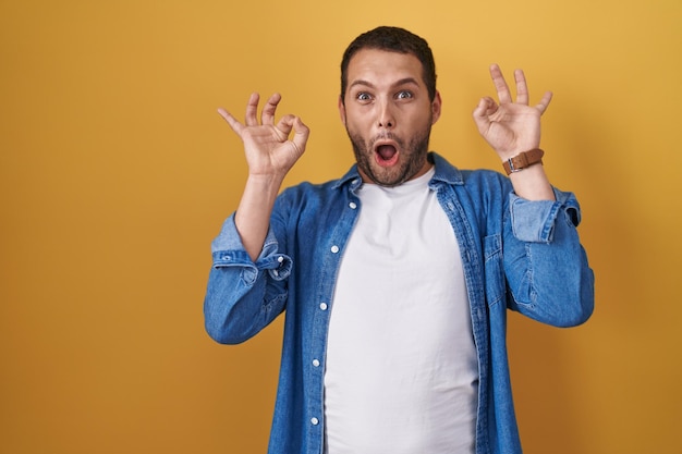 Hispanic man standing over yellow background looking surprised and shocked doing ok approval symbol with fingers. crazy expression