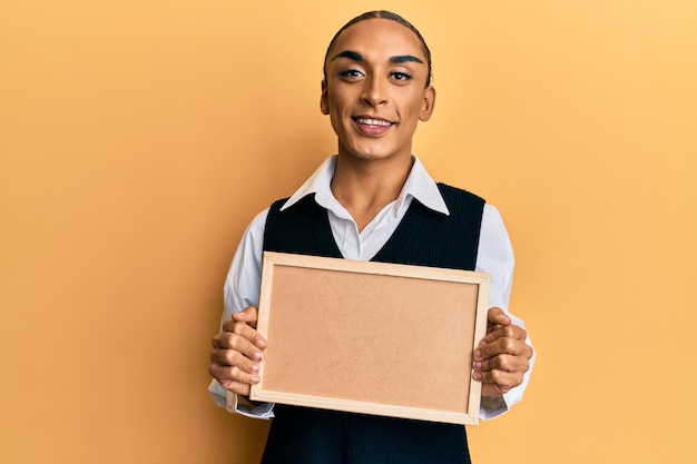 Free photo hispanic man wearing make up and long hair holding empty corkboard smiling with a happy and cool smile on face. showing teeth.