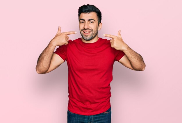 ひげを生やしたヒスパニック系の男性がカジュアルな赤い t シャツを着て、笑顔を見せ、指で歯と口を指しています。歯の健康の概念。