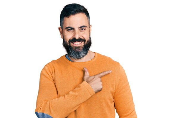 Латиноамериканец с бородой в повседневном зимнем свитере, веселый, с улыбкой на лице, указывающий рукой и пальцем в сторону со счастливым и естественным выражением лица