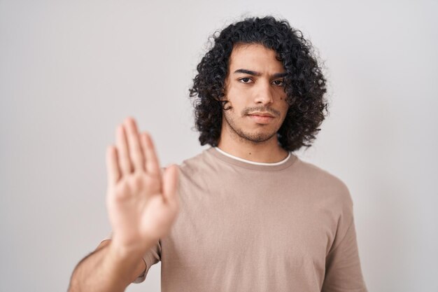 Hispanic man with curly hair standing over white background doing stop sing with palm of the hand warning expression with negative and serious gesture on the face