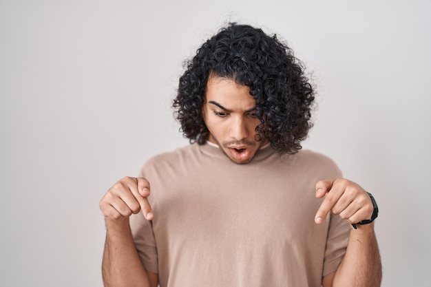 Uomo ispanico con i capelli ricci in piedi su sfondo bianco rivolto verso il basso con le dita che mostrano la faccia sorpresa della pubblicità e la bocca aperta