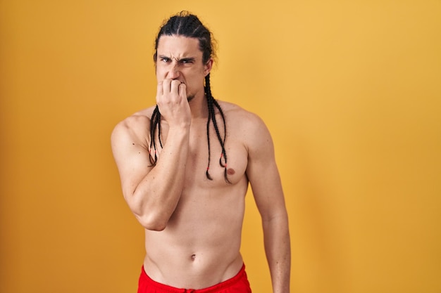 Hispanic man with long hair standing shirtless over yellow background looking stressed and nervous with hands on mouth biting nails anxiety problem