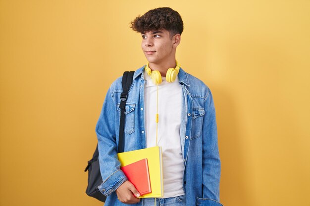 Hispanic teenager wearing student backpack and holding books smiling looking to the side and staring away thinking.