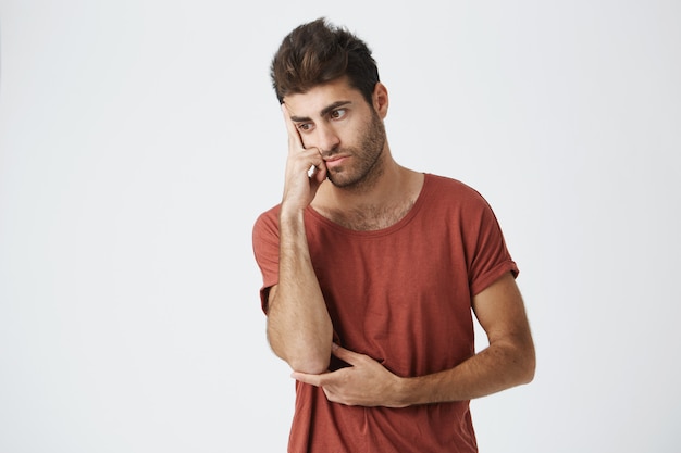 Hispanic unshaven good-looking guy in red t shirt grumpy looking aside holding hand on forehead upset about quarrel with friend.