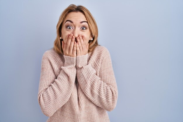 Hispanic woman standing over blue background laughing and embarrassed giggle covering mouth with hands, gossip and scandal concept