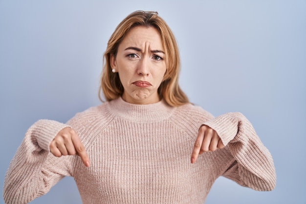 Free photo hispanic woman standing over blue background pointing down looking sad and upset, indicating direction with fingers, unhappy and depressed.