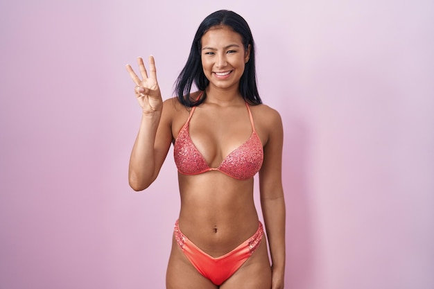 Foto gratuita donna ispanica che indossa bikini mostrando e puntando verso l'alto con le dita numero tre mentre sorride fiducioso e felice