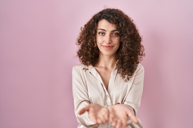 Hispanic woman with curly hair standing over pink background smiling with hands palms together receiving or giving gesture hold and protection