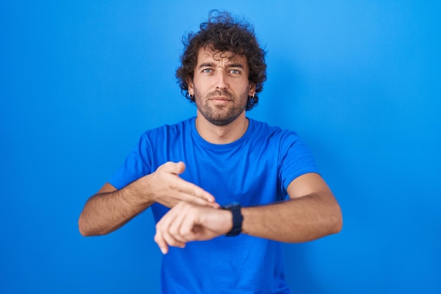 Испаноязычный молодой человек, стоящий на синем фоне в спешке, указывая на время просмотра, нетерпение, расстроен и зол из-за задержки крайнего срока