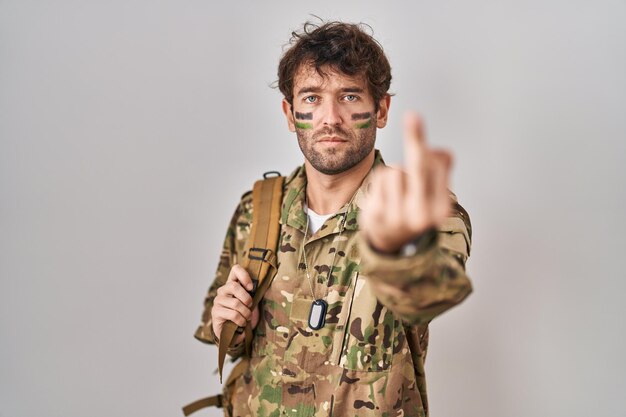 Hispanic young man wearing camouflage army uniform showing middle finger impolite and rude fuck off expression