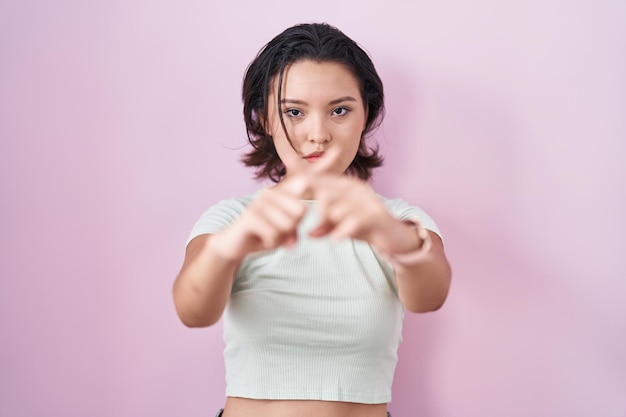 Hispanic young woman standing over pink background rejection expression crossing fingers doing negative sign