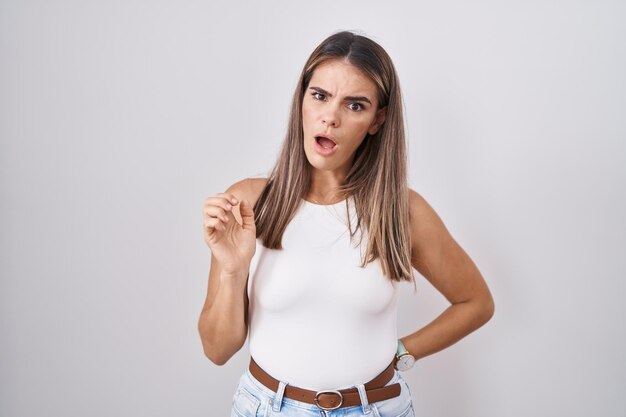 Hispanic young woman standing over white background in shock face, looking skeptical and sarcastic, surprised with open mouth