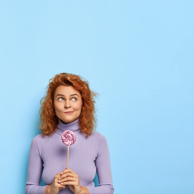 Free photo hmm can sugar make us happy? pensive young ginger woman stands with sweet lollipop in hands containing much sugar