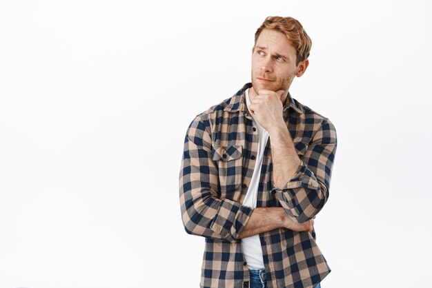 Hmm Handsome redhead man looking aside at advertisement and thinking deciding to buy something making choice standing thoughtful against white background