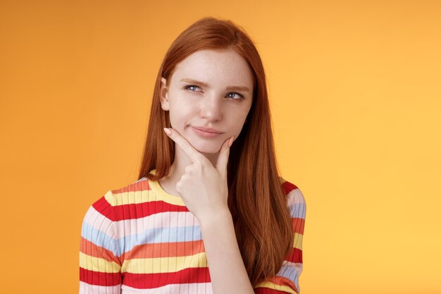 Hmm interesting. Suspicious doubtful attractive european redhead female pondering choice look hesitant thoughtful touch chin smirking tricky look upper left corner thinking, take decision.