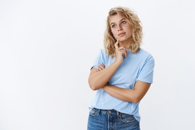 Free Photo hmm let me think. portrait of focused attractive female friend standing in thoughtful pose looking at upper left corner as remember, recalling thought touching jawline as daydreaming over white wall