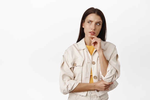 Hmm let me think. Thoughtful brunette woman furrows while ponder smth serious, looking at upper left corner pensive, making choice, standing  on white.