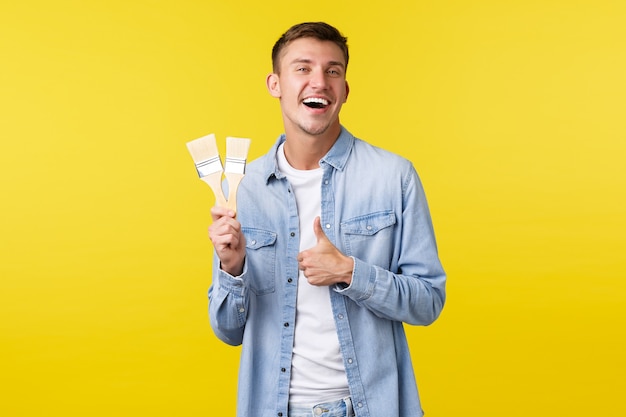 Free photo hobbies, leisure and people lifestyle concept. happy smiling satisfied guy showing thumbs-up and laughing joyfully as showing two painting brushes, standing yellow background, time for renovation.