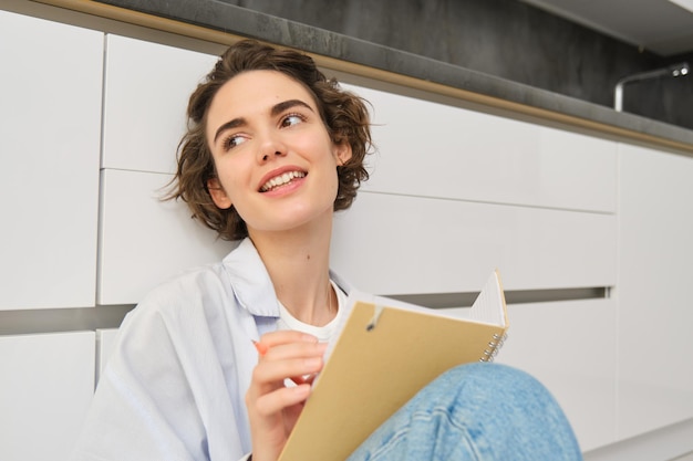 Free photo hobbies and lifestyle concept happy young woman sits on floor with pen and notebook wirtes in her