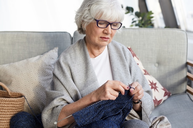 趣味、レジャー、退職の概念。針で灰色のソファに座って眼鏡をかけている格好良いエレガントな祖母は、彼女の孫のためにセーターを編んで、真剣に焦点を当てた表情をしています