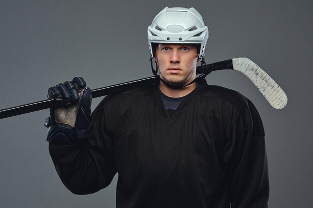 Hockey player wearing black protective gear and white helmet holds a hockey stick. Isolated on a gray background.