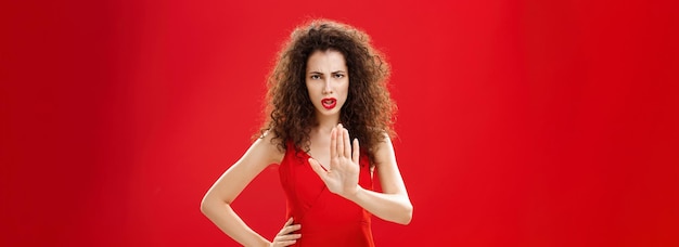Бесплатное фото Держись, стой, напряженная серьезно выглядящая недовольная взрослая женщина с вьющимися волосами в стильном красном платье